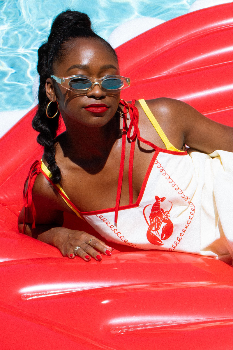 A woman lounging on a red inflatable pool float by a swimming pool, wearing a white one-piece swimsuit with red and yellow trim and a lobster graphic dress by Tuesday Bassen, along with cat-eye sunglasses.